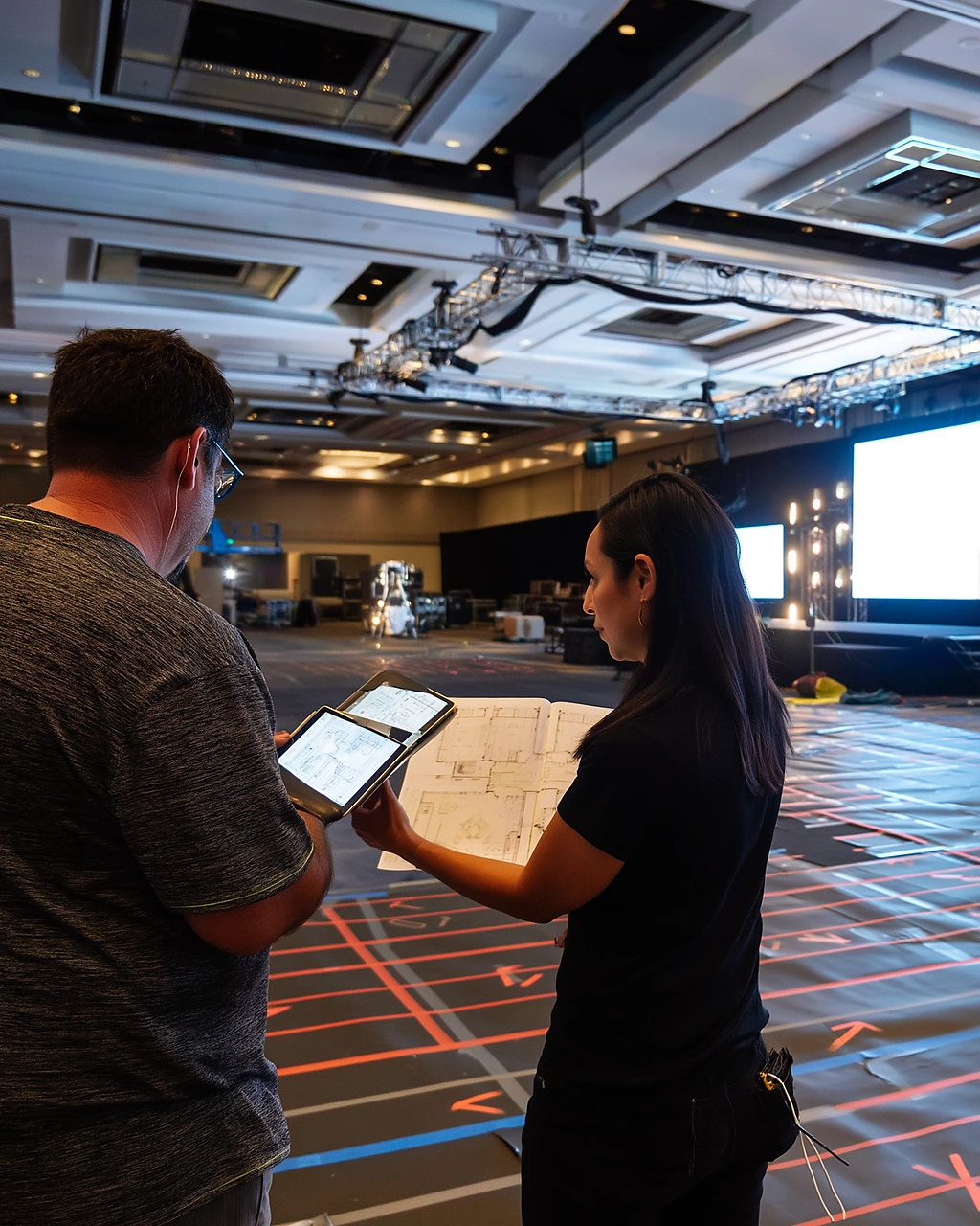 Event producers reviewing a floor plan and tablet on a ballroom floor before stage build is locked